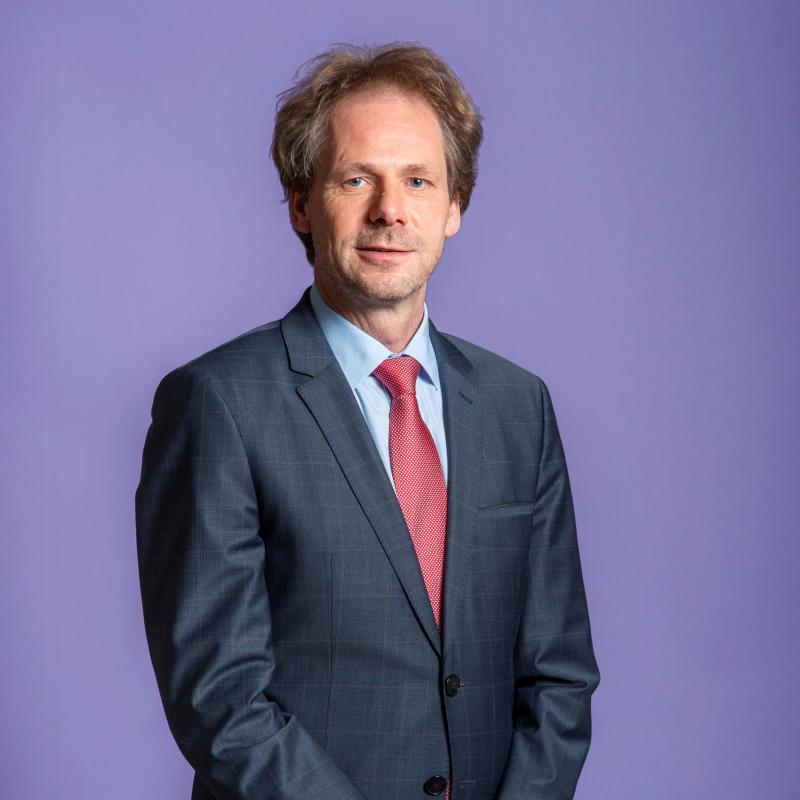 Foto van Bart Snels, een witte man. Hij draagt een zwart pak met een rode stropdas, heeft donkerblond haar en een vriendelijke uitdrukking op zijn gezicht.